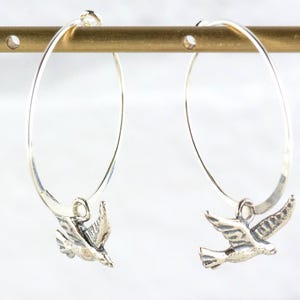 Peut inclure: Paire de boucles d'oreilles cr&eacute;oles en argent avec un charm en forme d'oiseau. Les boucles d'oreilles sont suspendues &agrave; une barre dor&eacute;e sur fond blanc. Les breloques oiseaux sont d&eacute;taill&eacute;es avec des plumes et des ailes.