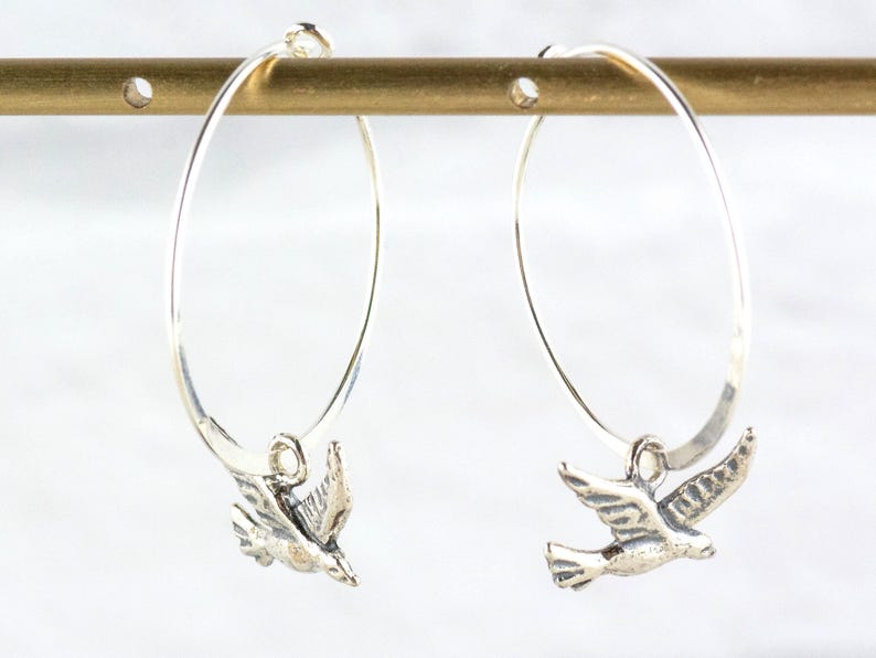 Peut inclure: Paire de boucles d'oreilles cr&eacute;oles en argent avec un charm en forme d'oiseau. Les boucles d'oreilles sont suspendues &agrave; une barre dor&eacute;e sur fond blanc. Les breloques oiseaux sont d&eacute;taill&eacute;es avec des plumes et des ailes.