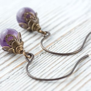 K&ouml;nnte beinhalten: Nahaufnahme von Amethyst-Ohrringen. Jeder Ohrring hat einen polierten, runden, lila Edelstein, der in einem verzierten, bronzefarbenen Metallgeh&auml;use gefasst ist. Die Ohrringe haben einen gebogenen Hakenverschluss. Der Hintergrund ist eine wei&szlig;e Holzoberfl&auml;che.