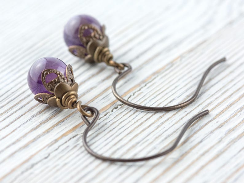 K&ouml;nnte beinhalten: Nahaufnahme von Amethyst-Ohrringen. Jeder Ohrring hat einen polierten, runden, lila Edelstein, der in einem verzierten, bronzefarbenen Metallgeh&auml;use gefasst ist. Die Ohrringe haben einen gebogenen Hakenverschluss. Der Hintergrund ist eine wei&szlig;e Holzoberfl&auml;che.