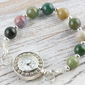 K&ouml;nnte beinhalten: Eine silberne Taschenuhr mit wei&szlig;em Zifferblatt und schwarzen Zeigern ist an einem Armband aus gr&uuml;nen, braunen und grauen Achatperlen befestigt.