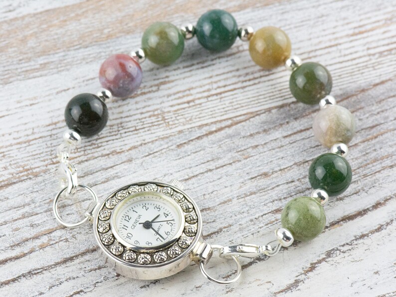 K&ouml;nnte beinhalten: Eine silberne Taschenuhr mit wei&szlig;em Zifferblatt und schwarzen Zeigern ist an einem Armband aus gr&uuml;nen, braunen und grauen Achatperlen befestigt.