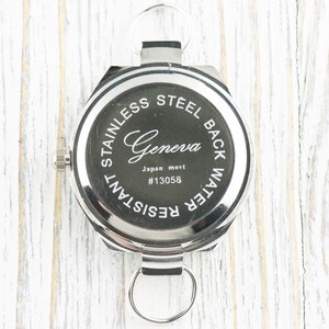 May include: The back of a silver watch with a black face. The watch has the words "Stainless Steel Back Water Resistant" and "Geneva Japan movt #13058" printed on the back.
