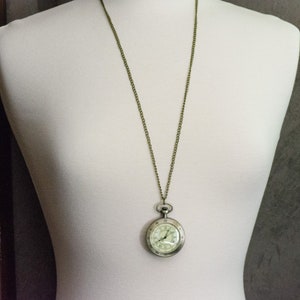 Op de afbeelding: Antieke zakhorloge hanger ketting. Het horloge heeft een zilverkleurige kast met een witte wijzerplaat en zwarte cijfers. De ketting is bronskleurig. Het horloge wordt getoond op een witte mannequin.