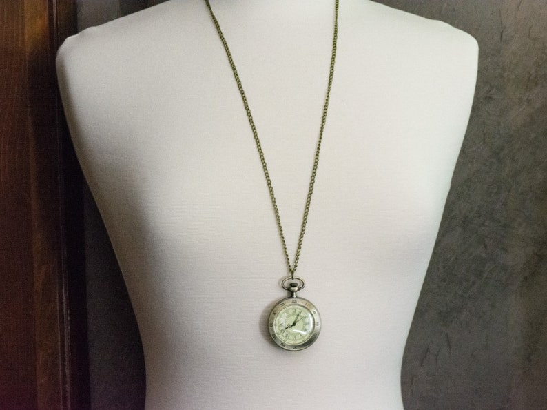 Op de afbeelding: Antieke zakhorloge hanger ketting. Het horloge heeft een zilverkleurige kast met een witte wijzerplaat en zwarte cijfers. De ketting is bronskleurig. Het horloge wordt getoond op een witte mannequin.