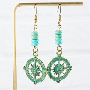 May include: A pair of turquoise and brass compass earrings. The earrings feature a compass design with a star in the centre and the letters N, S, E, and W around the edge. The compass is surrounded by turquoise beads.