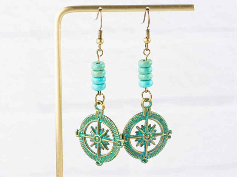 May include: A pair of turquoise and brass compass earrings. The earrings feature a compass design with a star in the centre and the letters N, S, E, and W around the edge. The compass is surrounded by turquoise beads.