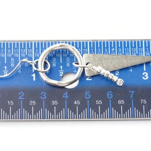 May include: A pair of silver earrings with a circle and a triangle pendant. The earrings are measured against a blue ruler, showing they are approximately 3 cm long.