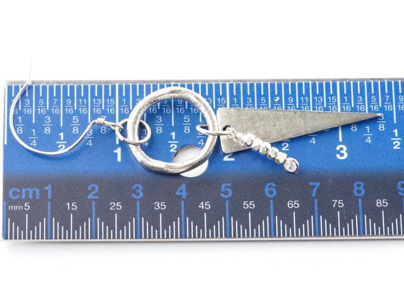 May include: A pair of silver earrings with a circle and a triangle pendant. The earrings are measured against a blue ruler, showing they are approximately 3 cm long.