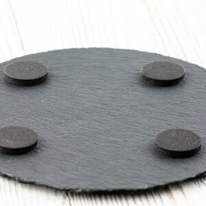 May include: A round, dark gray slate coaster with four small, round, black felt pads attached to the bottom. The coaster has a textured surface and a natural, organic shape.