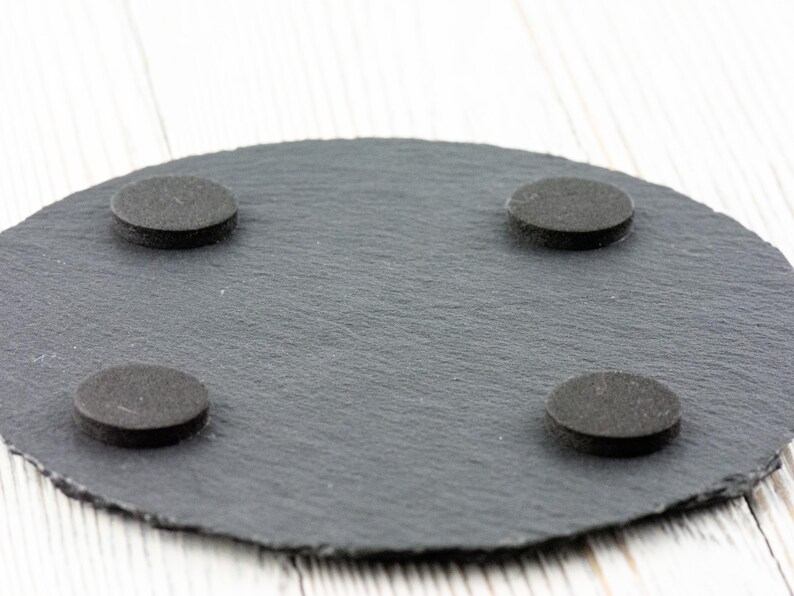 May include: A round, dark gray slate coaster with four small, round, black felt pads attached to the bottom. The coaster has a textured surface and a natural, organic shape.