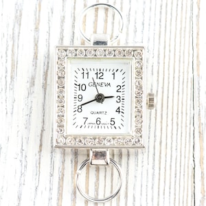May include: A silver-toned Geneva quartz watch with a square face, surrounded by rhinestones. The watch has a white dial with black numerals and hands. It features a ring at the top and bottom for attachment.