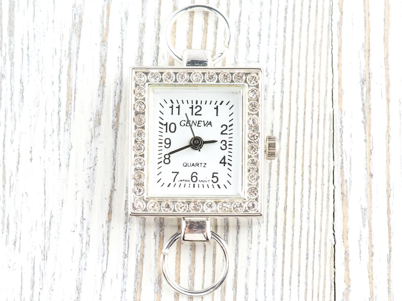 May include: A silver-toned Geneva quartz watch with a square face, surrounded by rhinestones. The watch has a white dial with black numerals and hands. It features a ring at the top and bottom for attachment.