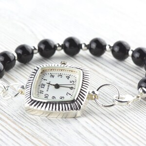 May include: A silver watch with a square face and a white dial, attached to a bracelet of large, round, black beads. The watch face has black numerals and hands. The bracelet is secured with a silver clasp.