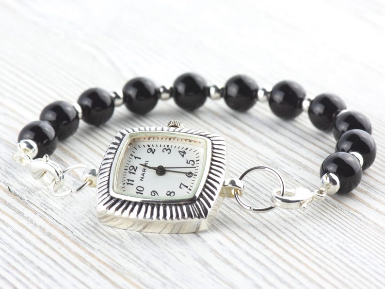May include: A silver watch with a square face and a white dial, attached to a bracelet of large, round, black beads. The watch face has black numerals and hands. The bracelet is secured with a silver clasp.