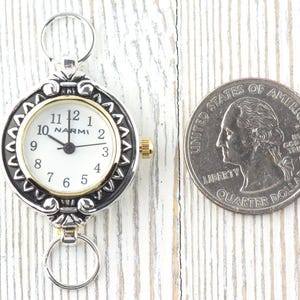 Pu&ograve; includere: Un orologio da tasca argentato e dorato con quadrante bianco e numeri neri. L'orologio ha un anello in alto e in basso per l'attacco. Una moneta da un quarto di dollaro &egrave; accanto all'orologio per il confronto delle dimensioni.