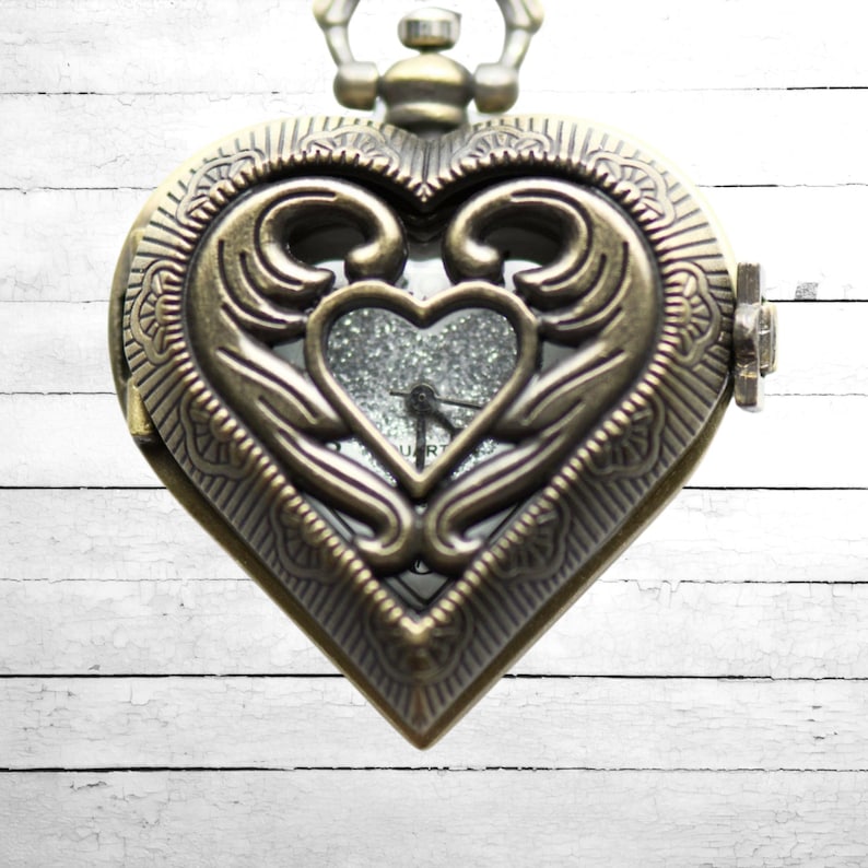 Puede incluir: Un reloj de bolsillo de estilo vintage con forma de coraz&oacute;n. El reloj tiene un acabado en bronce con detalles ornamentados y una peque&ntilde;a esfera en forma de coraz&oacute;n con purpurina. Las manecillas indican la hora.
