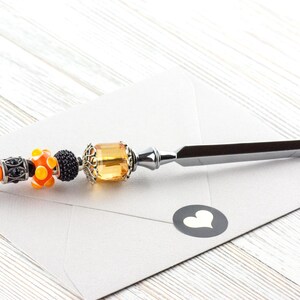 May include: Decorative letter opener with a silver blade and a beaded handle. The handle features orange, black, and yellow beads, along with a large amber-colored gem. The opener rests on a white envelope with a heart sticker.