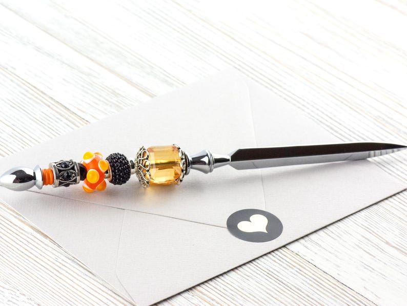 May include: Decorative letter opener with a silver blade and a beaded handle. The handle features orange, black, and yellow beads, along with a large amber-colored gem. The opener rests on a white envelope with a heart sticker.
