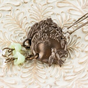May include: A bronze-toned hair accessory featuring a decorative circular element with floral and bird motifs. The hair pin has two tines and is adorned with light green and clear beads. The accessory is set against a white, textured background.