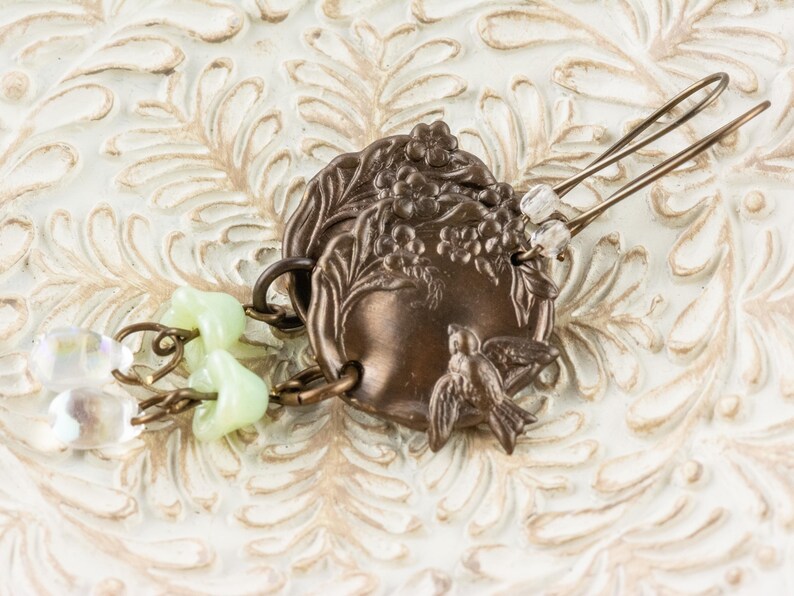 May include: A bronze-toned hair accessory featuring a decorative circular element with floral and bird motifs. The hair pin has two tines and is adorned with light green and clear beads. The accessory is set against a white, textured background.