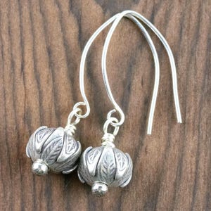 Pu&ograve; includere: Un paio di orecchini color argento con un design floreale. Gli orecchini hanno uno stile a goccia con un filo curvo che passa attraverso l'orecchio. Il design floreale &egrave; dettagliato e ha una piccola perla d'argento sul fondo. Lo sfondo &egrave; un legno scuro.
