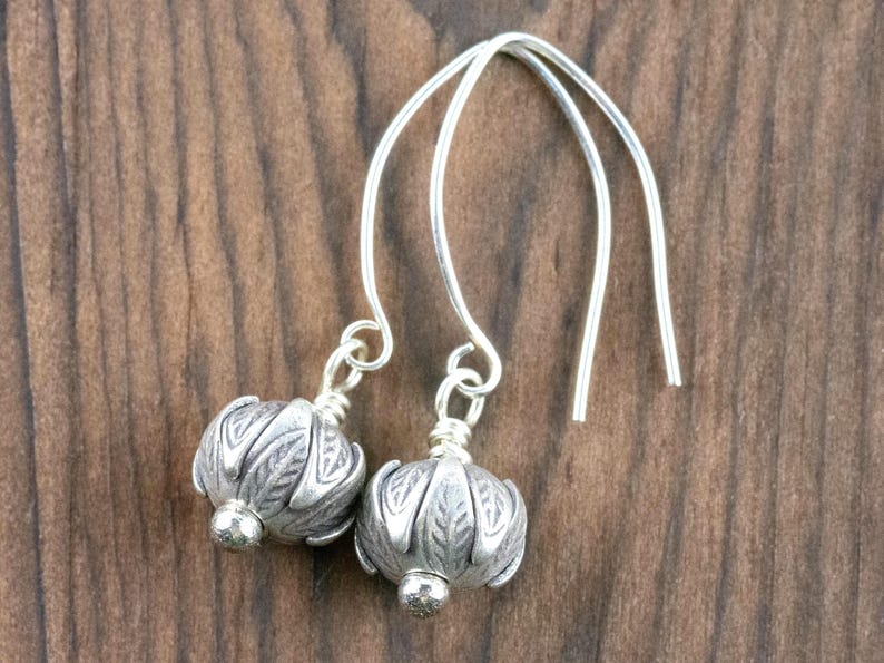 Pu&ograve; includere: Un paio di orecchini color argento con un design floreale. Gli orecchini hanno uno stile a goccia con un filo curvo che passa attraverso l'orecchio. Il design floreale &egrave; dettagliato e ha una piccola perla d'argento sul fondo. Lo sfondo &egrave; un legno scuro.