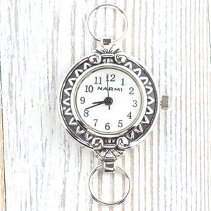 May include: Silver-toned pocket watch with a white face and black numerals. The watch has a decorative bezel and two rings for attachment. The brand name "NARMI" is printed on the watch face.