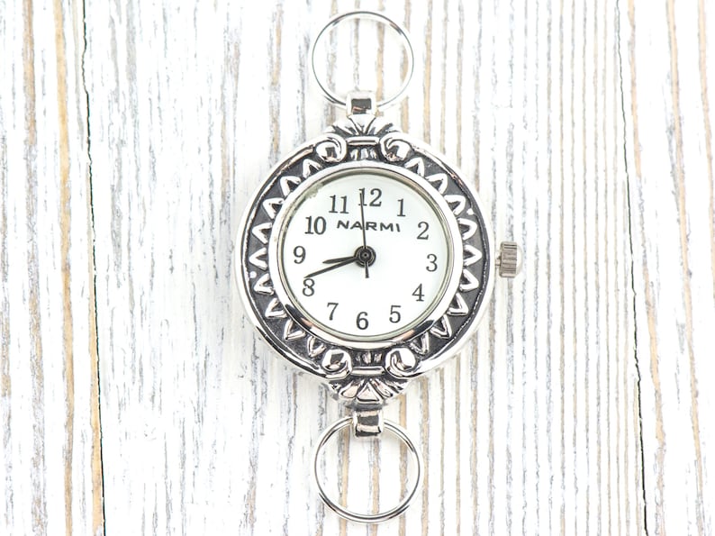 Op de afbeelding: Zilverkleurig zakhorloge met een witte wijzerplaat en zwarte cijfers. Het horloge heeft een decoratieve rand en twee ringen voor bevestiging. De merknaam "NARMI" staat op de wijzerplaat.
