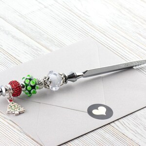 May include: A silver letter opener with decorative beads and a Christmas tree charm. The beads are red, green, and clear, and the opener rests on a gray envelope with a heart sticker. The opener is approximately 8 inches long.