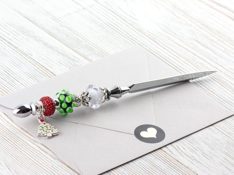 May include: A silver letter opener with decorative beads and a Christmas tree charm. The beads are red, green, and clear, and the opener rests on a gray envelope with a heart sticker. The opener is approximately 8 inches long.