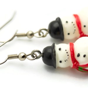 May include: A pair of silver snowman dangle earrings. Each snowman is white with a black top hat, red scarf, and black eyes and buttons.