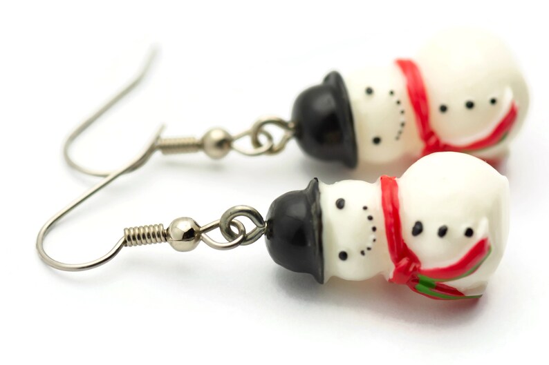 May include: A pair of silver snowman dangle earrings. Each snowman is white with a black top hat, red scarf, and black eyes and buttons.
