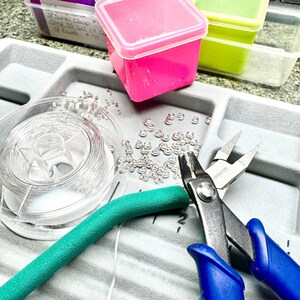 May include: A jewelry-making workspace with various supplies. Includes a spool of clear wire, small silver jump rings, a teal foam tube, and blue-handled pliers. Pink and green plastic containers are also visible, along with handwritten labels.