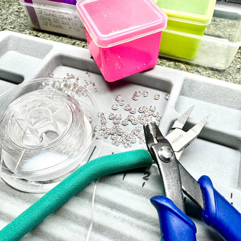May include: A jewelry-making workspace with various supplies. Includes a spool of clear wire, small silver jump rings, a teal foam tube, and blue-handled pliers. Pink and green plastic containers are also visible, along with handwritten labels.