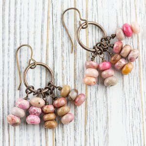 Op de afbeelding: Handgemaakte oorbellen met bronzen ringen en strengen ovale kralen in roze, perzik en bruin. De oorbellen worden gepresenteerd op een wit houten oppervlak. Een uniek accessoire met kralen in verschillende kleuren.