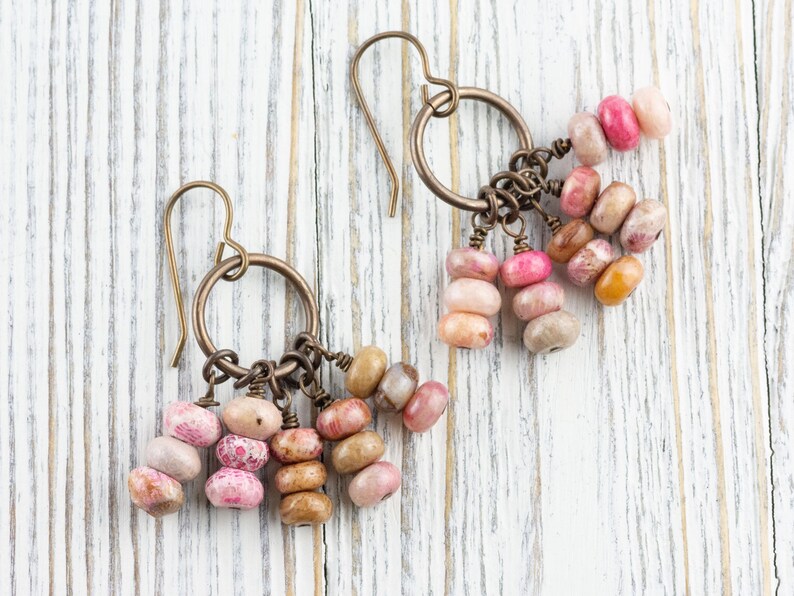 Op de afbeelding: Handgemaakte oorbellen met bronzen ringen en strengen ovale kralen in roze, perzik en bruin. De oorbellen worden gepresenteerd op een wit houten oppervlak. Een uniek accessoire met kralen in verschillende kleuren.