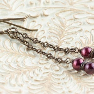 Puede incluir: Pendientes colgantes con est&eacute;tica vintage. Los pendientes presentan un dise&ntilde;o de cadena con peque&ntilde;as cuentas redondas de color p&uacute;rpura oscuro. El metal tiene un acabado bronce. Los pendientes se exhiben sobre una superficie blanca texturizada.