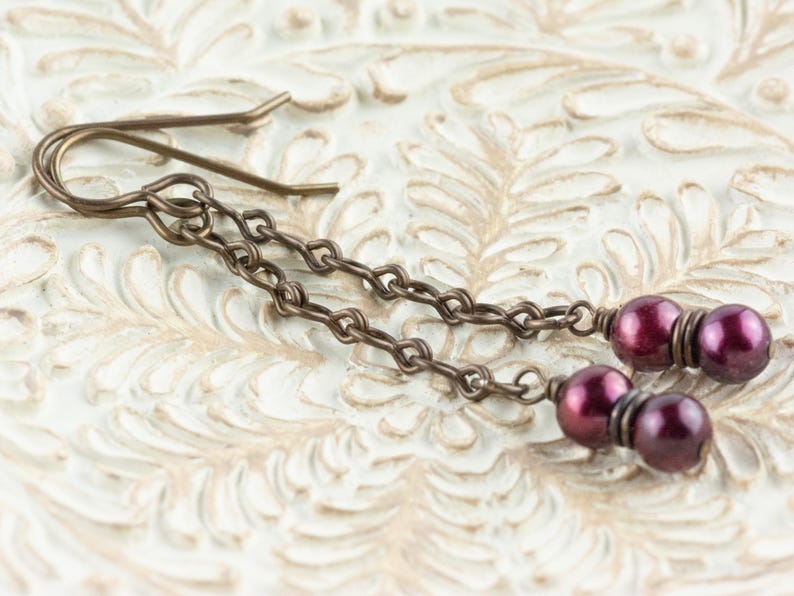 Puede incluir: Pendientes colgantes con est&eacute;tica vintage. Los pendientes presentan un dise&ntilde;o de cadena con peque&ntilde;as cuentas redondas de color p&uacute;rpura oscuro. El metal tiene un acabado bronce. Los pendientes se exhiben sobre una superficie blanca texturizada.
