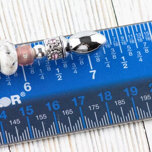May include: A close-up of a silver charm bracelet with various beads, including a silver bead with a textured surface, a white bead with black speckles, a pink bead, and a silver bead with a small pink flower design. The bracelet is measured on a blue ruler with black markings, showing a length of 7 inches.