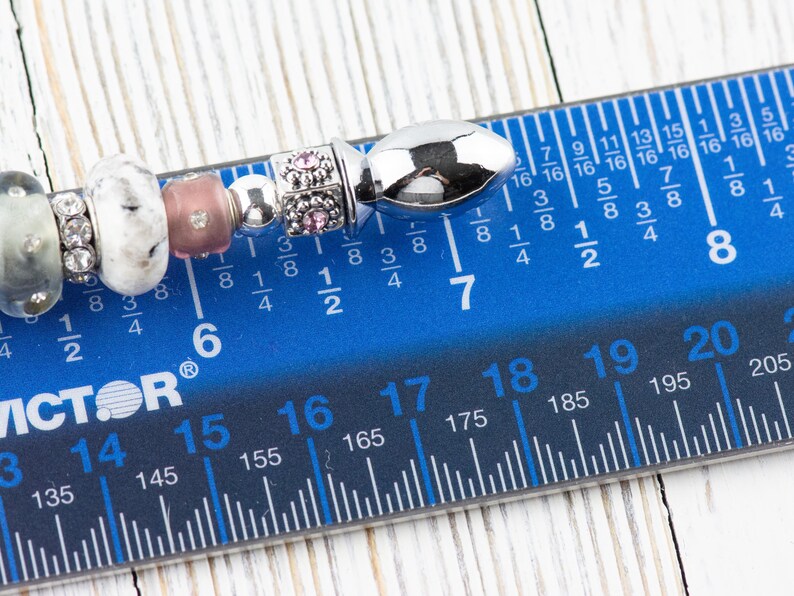 May include: A close-up of a silver charm bracelet with various beads, including a silver bead with a textured surface, a white bead with black speckles, a pink bead, and a silver bead with a small pink flower design. The bracelet is measured on a blue ruler with black markings, showing a length of 7 inches.