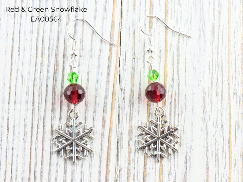 Pode incluir: Brincos de floco de neve prateados com detalhes em vermelho e verde. Cada brinco apresenta um pingente de floco de neve prateado, uma conta vermelha facetada e uma conta verde. O texto "Red & Green Snowflake EA00564" &eacute; vis&iacute;vel.
