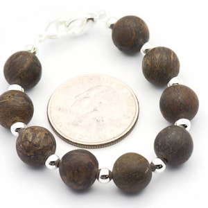 Puede incluir: Un brazalete de cuentas de madera marr&oacute;n con cuentas espaciadoras plateadas. El brazalete es aproximadamente del tama&ntilde;o de una moneda de 25 c&eacute;ntimos.