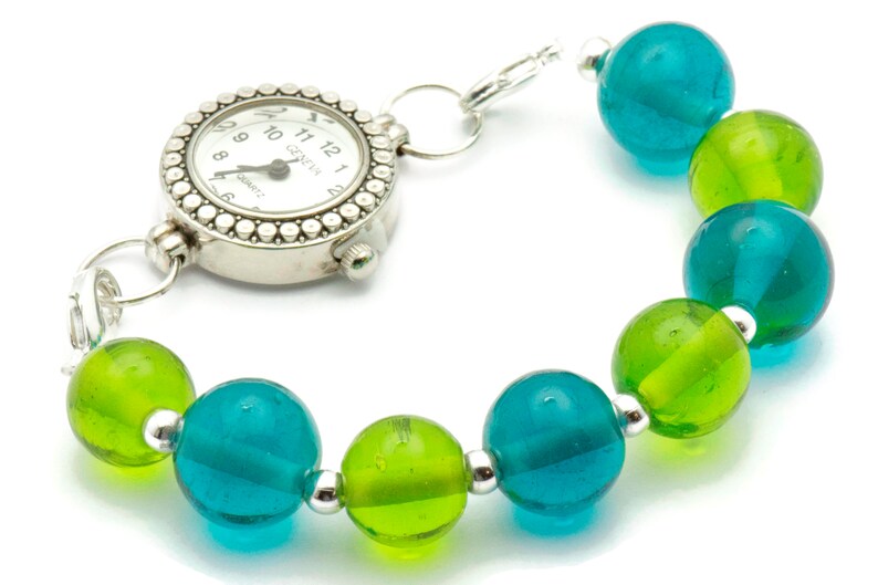Puede incluir: Un reloj plateado con una esfera blanca y agujas negras unidas a una pulsera hecha de cuentas de vidrio turquesa y verde alternas.