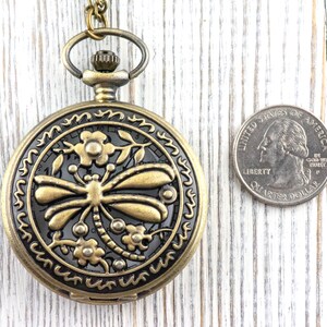 May include: Antique brass pocket watch with a dragonfly design. The watch has a detailed floral border and a black background. A US quarter is next to the watch for size comparison.