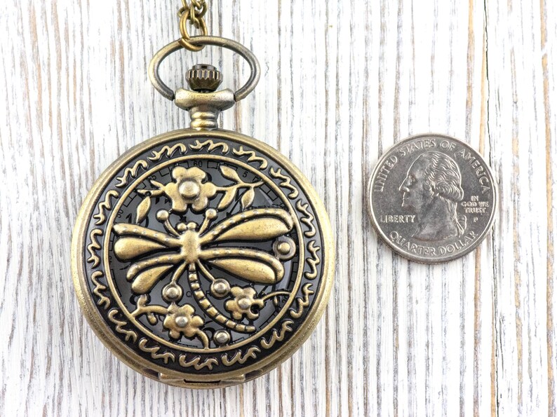 May include: Antique brass pocket watch with a dragonfly design. The watch has a detailed floral border and a black background. A US quarter is next to the watch for size comparison.