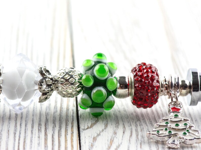 May include: A close-up of a charm bracelet with various beads and charms. The bracelet features a white faceted bead, a silver filigree bead, a green glass bead, and a red, textured bead. A silver Christmas tree charm hangs from the bracelet.