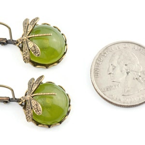 May include: A pair of green and gold earrings featuring a dragonfly design. The earrings are made with a round green stone and a gold-toned dragonfly charm. The earrings are shown next to a US quarter for size reference.