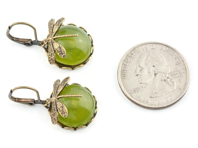May include: A pair of green and gold earrings featuring a dragonfly design. The earrings are made with a round green stone and a gold-toned dragonfly charm. The earrings are shown next to a US quarter for size reference.