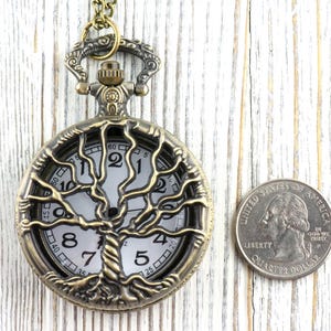 May include: Vintage bronze pocket watch featuring a tree of life motif. The watch face is white with black numerals and hands. A quarter coin is next to the watch for size comparison. The watch's diameter is approximately 4 cm.